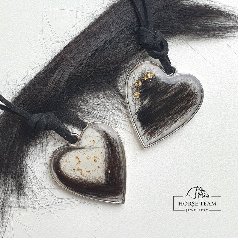 May include: Two silver heart-shaped pendants with black horse hair encased in clear resin. Each pendant has gold flakes embedded in the resin. The pendants are attached to black cords.