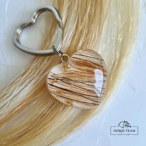 May include: A heart-shaped keyring with a silver key ring. The heart is filled with brown horse hair and gold flakes. The keyring is on a background of blonde horse hair.