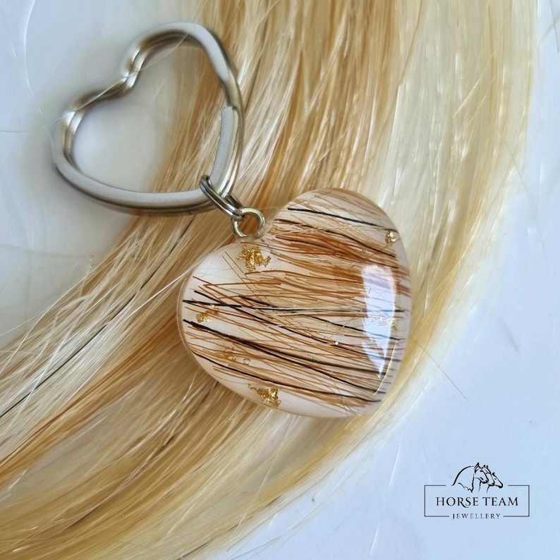 May include: A heart-shaped keyring with a silver key ring. The heart is filled with brown horse hair and gold flakes. The keyring is on a background of blonde horse hair.