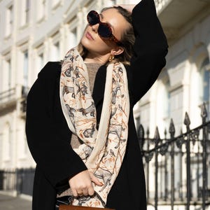 Può includere: Una donna indossa un cappotto nero, occhiali da sole e una sciarpa fantasia, tenendo in mano una borsa marrone con texture di coccodrillo. La sciarpa presenta un'illustrazione ripetuta di cani. Lo sfondo include un edificio bianco e una recinzione in ferro battuto nero.