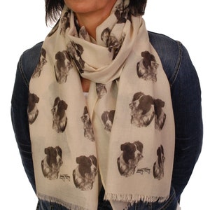 May include: A cream-colored scarf with a repeating pattern of black and white border collie dog faces. The scarf is made of a soft, lightweight fabric and has fringed edges.