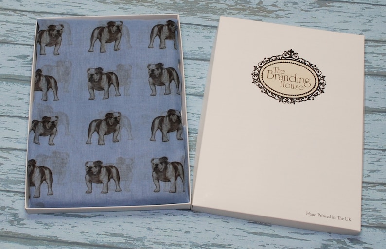 May include: A blue silk scarf with a repeating pattern of brown bulldogs. The scarf is packaged in a white box with the text "The Branding House" and "Hand Printed in the UK".