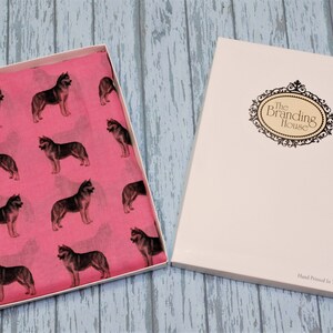 May include: A bright pink scarf featuring a repeating pattern of dark grey husky dogs, presented in an open white gift box. The box lid displays an ornate black oval frame with the text The Branding House and Hand Printed in The UK.