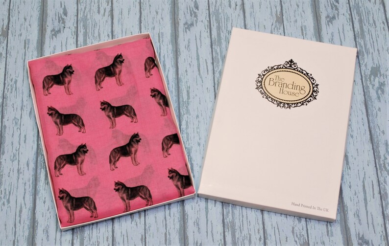 May include: A bright pink scarf featuring a repeating pattern of dark grey husky dogs, presented in an open white gift box. The box lid displays an ornate black oval frame with the text The Branding House and Hand Printed in The UK.