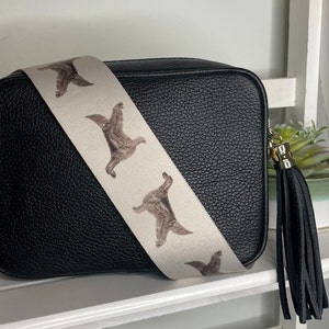 K&ouml;nnte beinhalten: Schwarze Leder-Umh&auml;ngetasche mit einem wei&szlig;en Riemen, der ein sich wiederholendes Muster aus braunen Hunden zeigt. Die Tasche hat einen schwarzen Quasten.