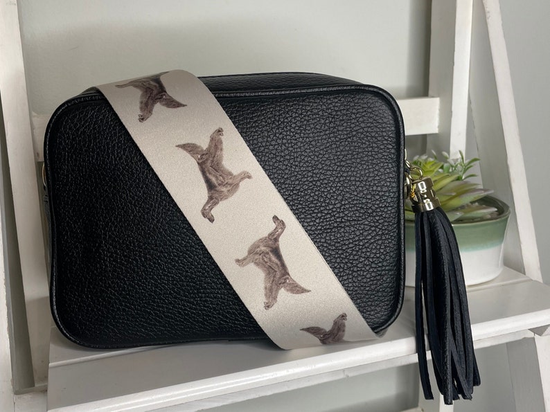 K&ouml;nnte beinhalten: Schwarze Leder-Umh&auml;ngetasche mit einem wei&szlig;en Riemen, der ein sich wiederholendes Muster aus braunen Hunden zeigt. Die Tasche hat einen schwarzen Quasten.