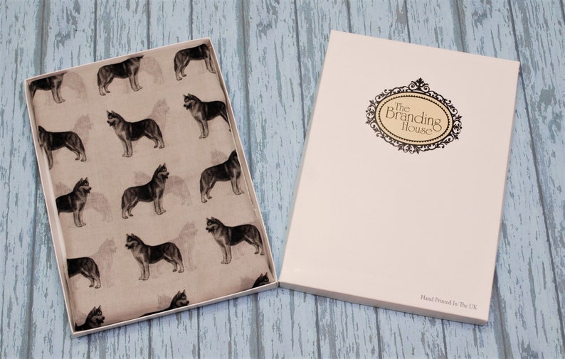 May include: A rectangular gift box containing a light beige fabric featuring a repeating pattern of dark grey husky dogs. The box lid is white with a decorative black oval frame containing the text The Branding House and Hand Printed In The UK.