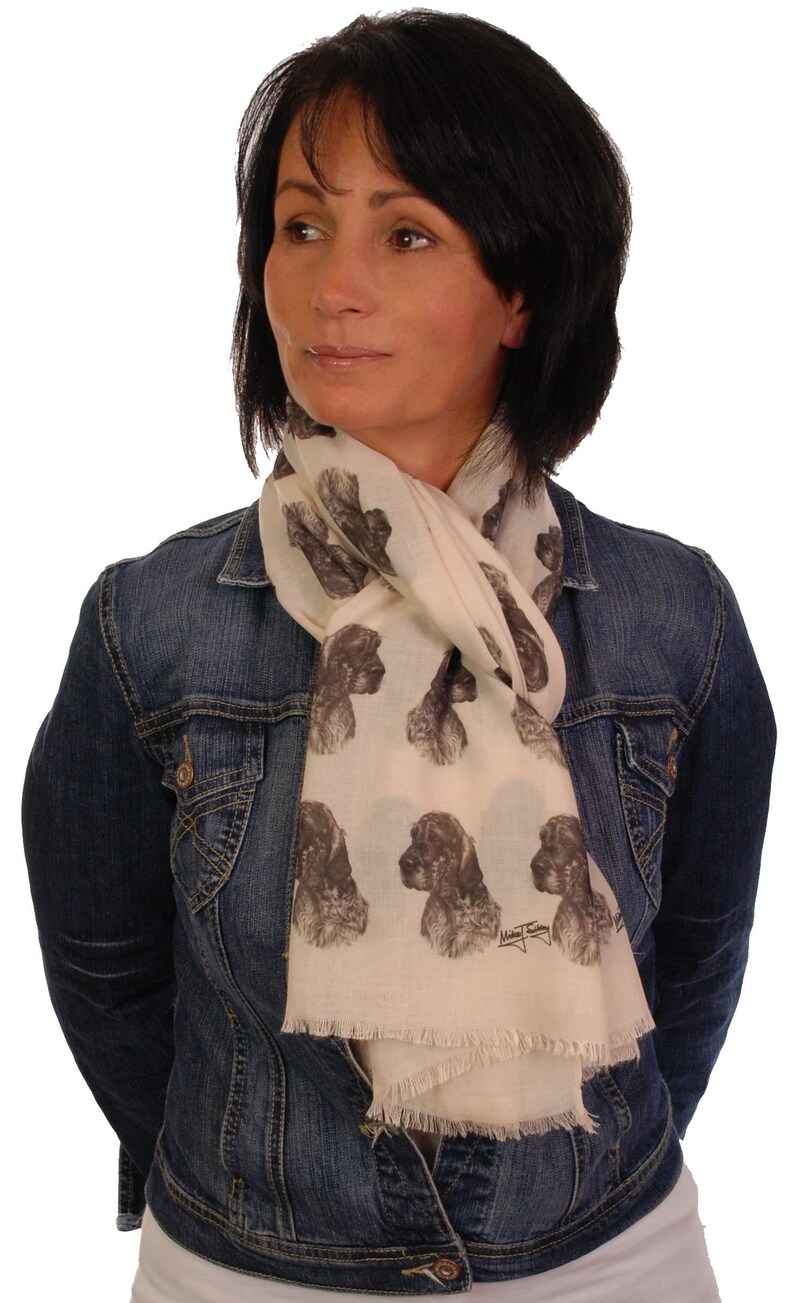 May include: A woman wearing a denim jacket over a white scarf with a brown and white dog print. The scarf has a fringe edge and the dog print is repeated throughout the scarf.