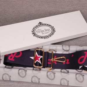 May include: A navy blue and red guitar strap with a gold star and buckle. The strap is packaged in a white box with the text "The Branding House".