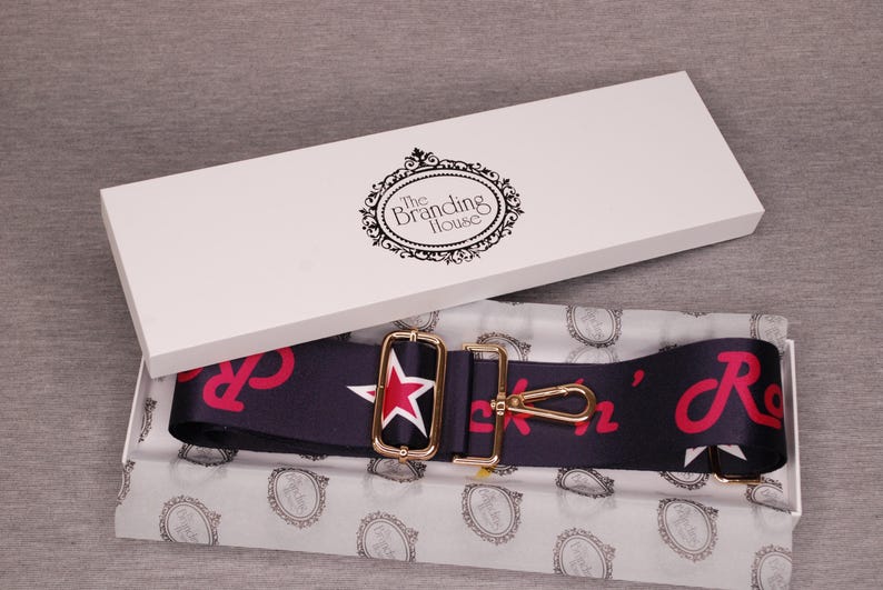 May include: A navy blue and red guitar strap with a gold star and buckle. The strap is packaged in a white box with the text "The Branding House".