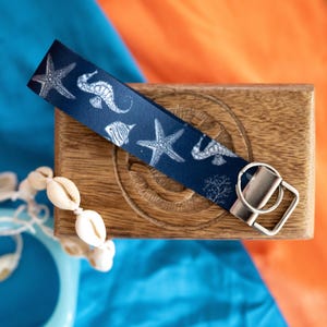 May include: A navy blue key fob wrist strap with a white nautical design of starfish, seahorses, and fish. The key fob has a silver metal ring and is on a wooden box. Seashells are in the foreground.