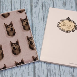 Pu&ograve; includere: Una scatola regalo rettangolare bianca con un coperchio che riporta il testo The Branding House e Hand Printed In The UK. All'interno della scatola aperta si trova un tessuto rosa chiaro con un motivo ripetuto di volti di cani husky dal pelo grigio e bianco.