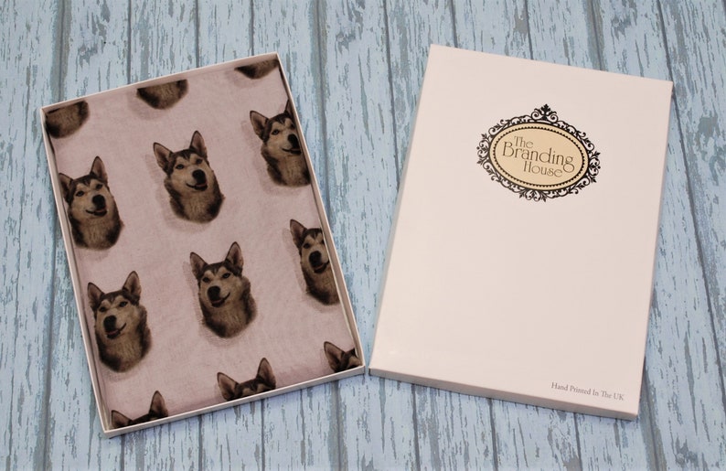 Pu&ograve; includere: Una scatola regalo rettangolare bianca con un coperchio che riporta il testo The Branding House e Hand Printed In The UK. All'interno della scatola aperta si trova un tessuto rosa chiaro con un motivo ripetuto di volti di cani husky dal pelo grigio e bianco.