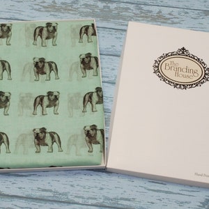 May include: A green fabric scarf with a repeating pattern of brown and white bulldogs. The scarf is in a white box with the text "The Branding House" and "Hand Printed in the UK".