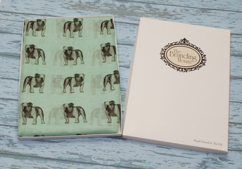 May include: A green fabric scarf with a repeating pattern of brown and white bulldogs. The scarf is in a white box with the text "The Branding House" and "Hand Printed in the UK".