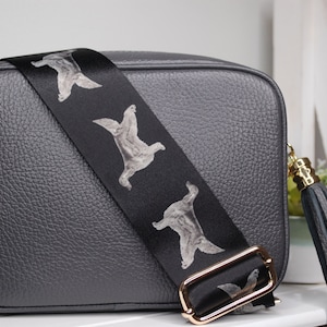 K&ouml;nnte beinhalten: Eine graue Leder-Umh&auml;ngetasche mit einem schwarzen verstellbaren Riemen, der ein sich wiederholendes Muster aus wei&szlig;en Hunden zeigt. Die Tasche hat eine goldfarbene Schnalle und einen schwarzen Quasten.
