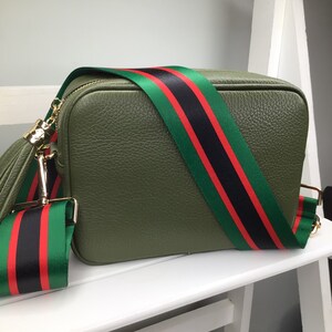 May include: Olive green crossbody bag with a textured finish. The bag features a wide strap with green, black, and red stripes. Gold-tone hardware and a matching tassel add detail. The bag is sitting on a white surface.