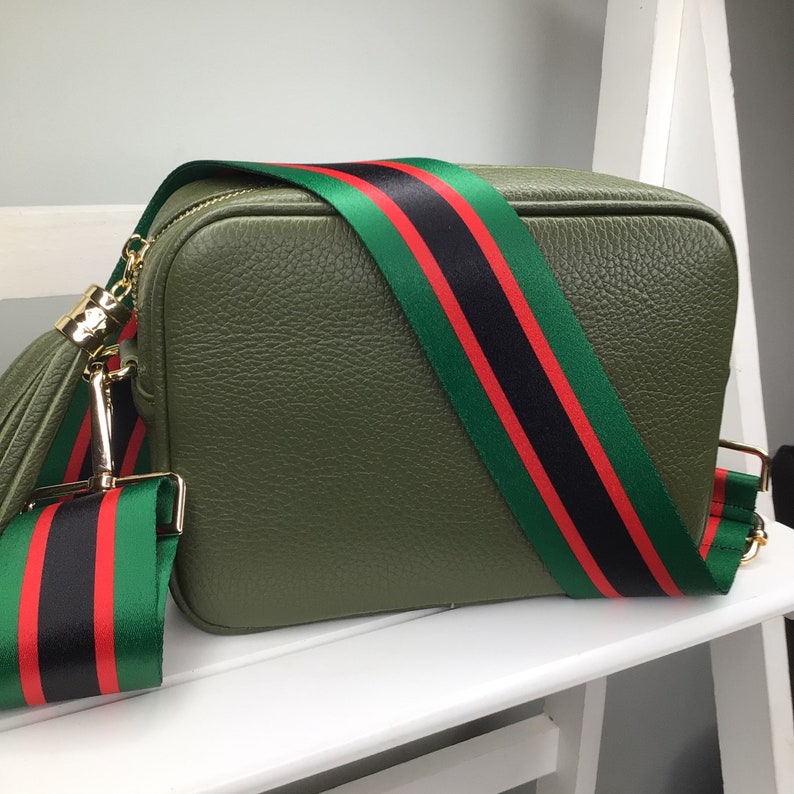 May include: Olive green crossbody bag with a textured finish. The bag features a wide strap with green, black, and red stripes. Gold-tone hardware and a matching tassel add detail. The bag is sitting on a white surface.