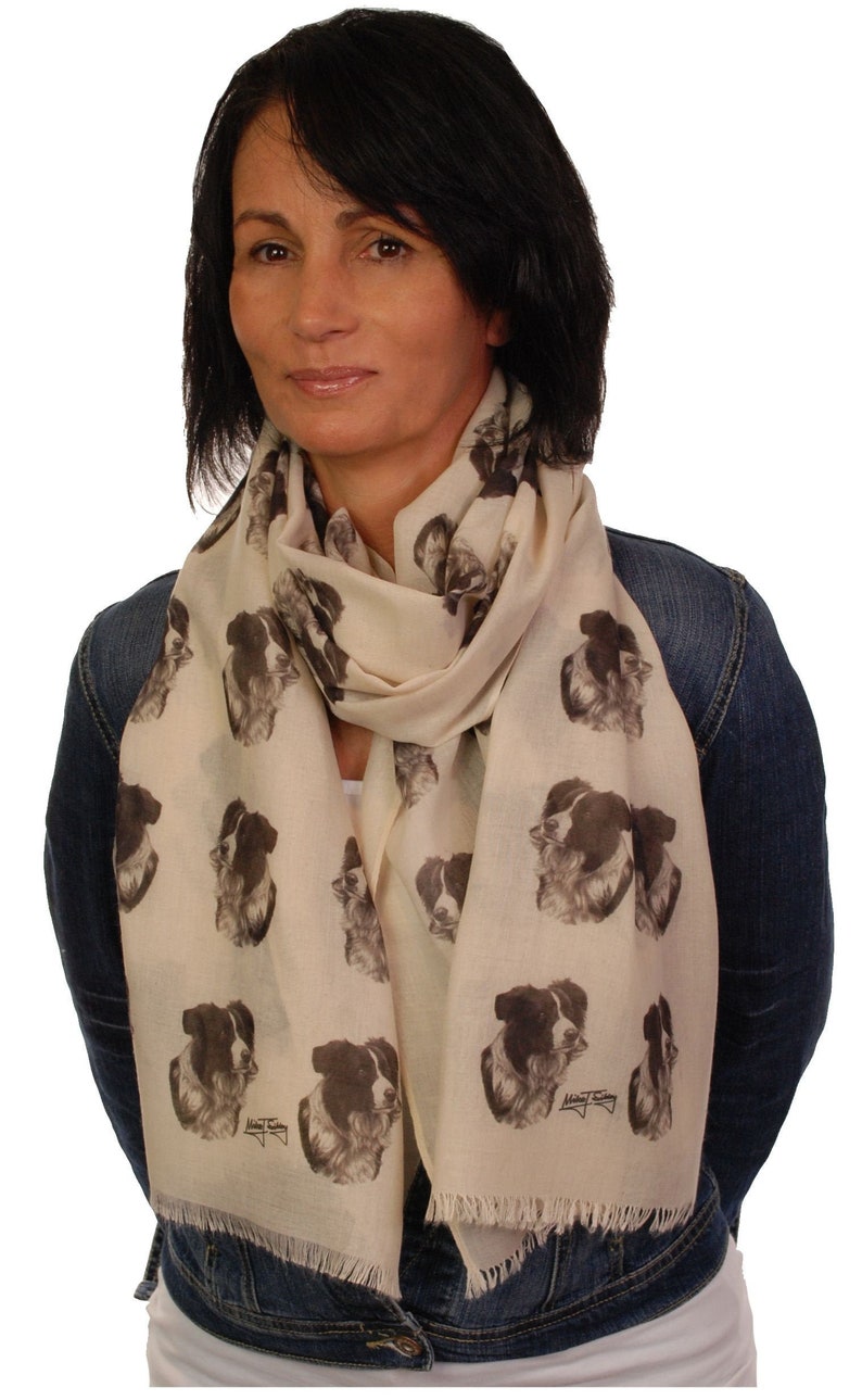May include: A cream-colored scarf with a repeating pattern of black and white border collie dog faces. The scarf is made of a soft, lightweight fabric and has fringed edges.