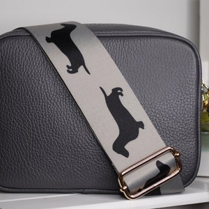 May include: A gray leather camera bag with a detachable strap featuring a repeating pattern of black dachshund silhouettes. The strap has a gold metal buckle and the bag has a black tassel.