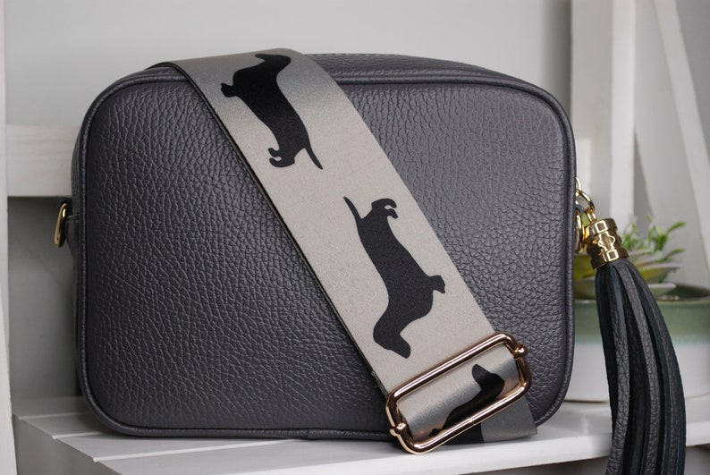 May include: A gray leather camera bag with a detachable strap featuring a repeating pattern of black dachshund silhouettes. The strap has a gold metal buckle and the bag has a black tassel.