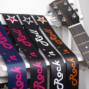 Puede incluir: Cinco correas de guitarra con estrellas de diferentes colores y el texto "Rock 'n' Roll" impreso en ellas. Las correas son negras, azul marino y blancas con letras rosas, naranjas y blancas.