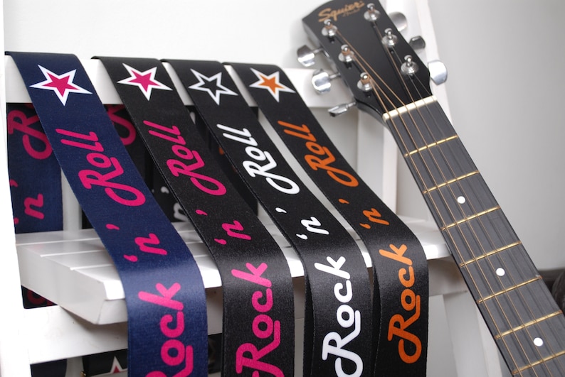 May include: Five guitar straps with different colored stars and the text "Rock 'n' Roll" printed on them. The straps are black, navy blue, and white with pink, orange, and white lettering.