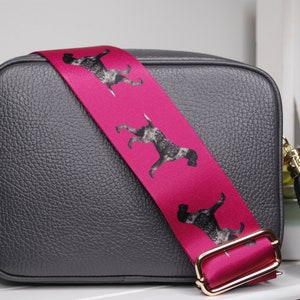 May include: A gray leather crossbody bag with a pink adjustable strap featuring a repeating pattern of black and white dogs. The strap has a gold buckle and the bag has a black tassel.