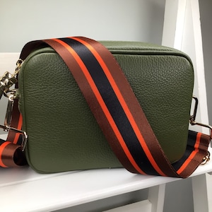 May include: Olive green crossbody bag with a wide, striped strap in brown, black, and orange. The bag has a rectangular shape and gold-colored hardware. The strap is adjustable and appears to be made of a durable material.