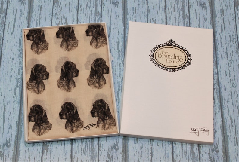 May include: A beige scarf with a repeating pattern of black and white dog illustrations. The scarf is in a white box with a label that says "The Branding House".