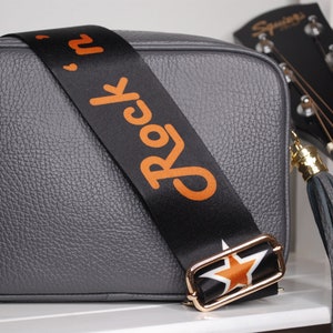 May include: Gray leather crossbody bag with a black and orange adjustable strap that says "Rock 'n' Roll" and a gold star buckle. The bag has a gray leather tassel.