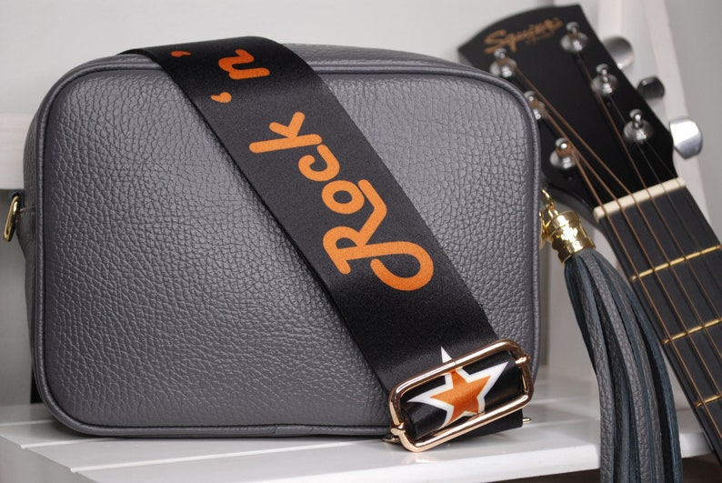 May include: Gray leather crossbody bag with a black and orange adjustable strap that says "Rock 'n' Roll" and a gold star buckle. The bag has a gray leather tassel.