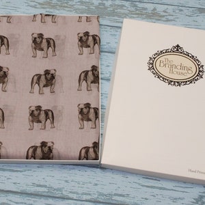 May include: A light pink scarf with a repeating pattern of brown bulldog illustrations. The scarf is packaged in a white box with the text "The Branding House" printed on the front. The box also has the text "Hand Printed in the U.K." printed on the bottom.
