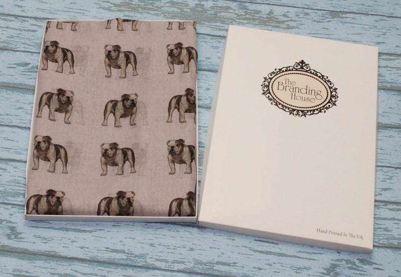 May include: A light pink scarf with a repeating pattern of brown bulldog illustrations. The scarf is packaged in a white box with the text "The Branding House" printed on the front. The box also has the text "Hand Printed in the U.K." printed on the bottom.
