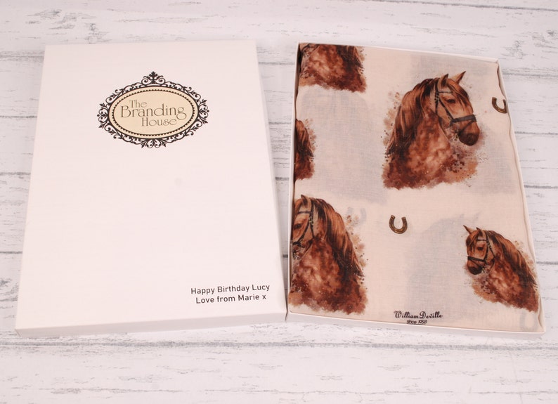 Puede incluir: Una caja de regalo blanca con una bufanda de estampado de caballo marr&oacute;n en su interior. La caja tiene un logotipo que dice "The Branding House" y un mensaje que dice "Happy Birthday Lucy Love from Marie x". La bufanda tiene un estampado de caballo marr&oacute;n con un dise&ntilde;o de herradura.