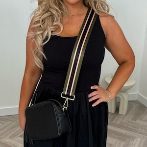May include: Black crossbody bag with a textured finish and a decorative tassel. The bag features a wide strap with olive green and pink stripes. The strap is attached to the bag with gold-colored hardware. The bag is worn over a black dress.