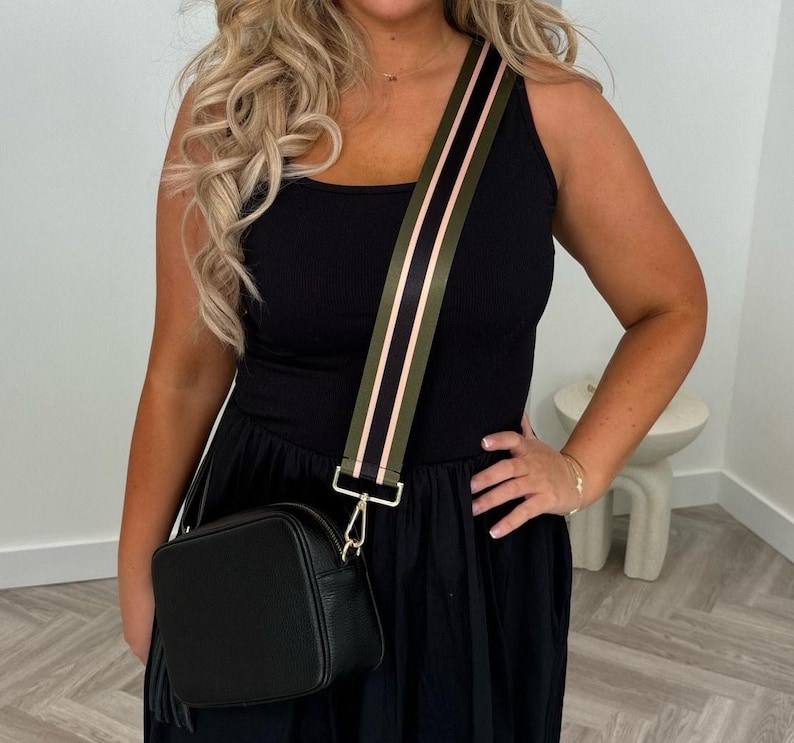 May include: Black crossbody bag with a textured finish and a decorative tassel. The bag features a wide strap with olive green and pink stripes. The strap is attached to the bag with gold-colored hardware. The bag is worn over a black dress.