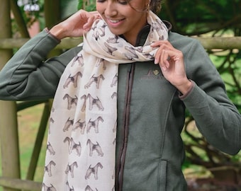 May include: A cream-colored scarf featuring a repeating pattern of brown and black Airedale Terrier dog silhouettes. The scarf is worn over a dark olive green zip-up fleece jacket with a small embroidered logo on the upper left chest area.