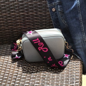 May include: A gray leather crossbody bag with a black and white adjustable strap. The strap has pink stars and the text "Rock 'n' Roll" printed on it.