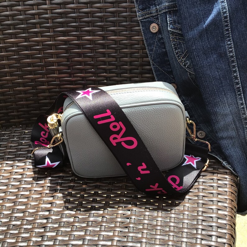 May include: A gray leather crossbody bag with a black and white adjustable strap. The strap has pink stars and the text "Rock 'n' Roll" printed on it.