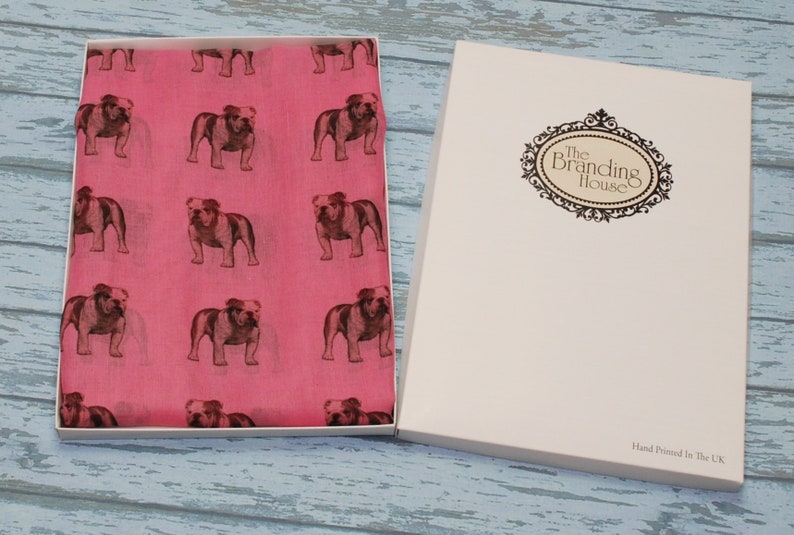 May include: A pink scarf with a repeating pattern of brown bulldog silhouettes. The scarf is in a white box with the text "The Branding House" on the lid. The bottom of the box says "Hand Printed in the UK."