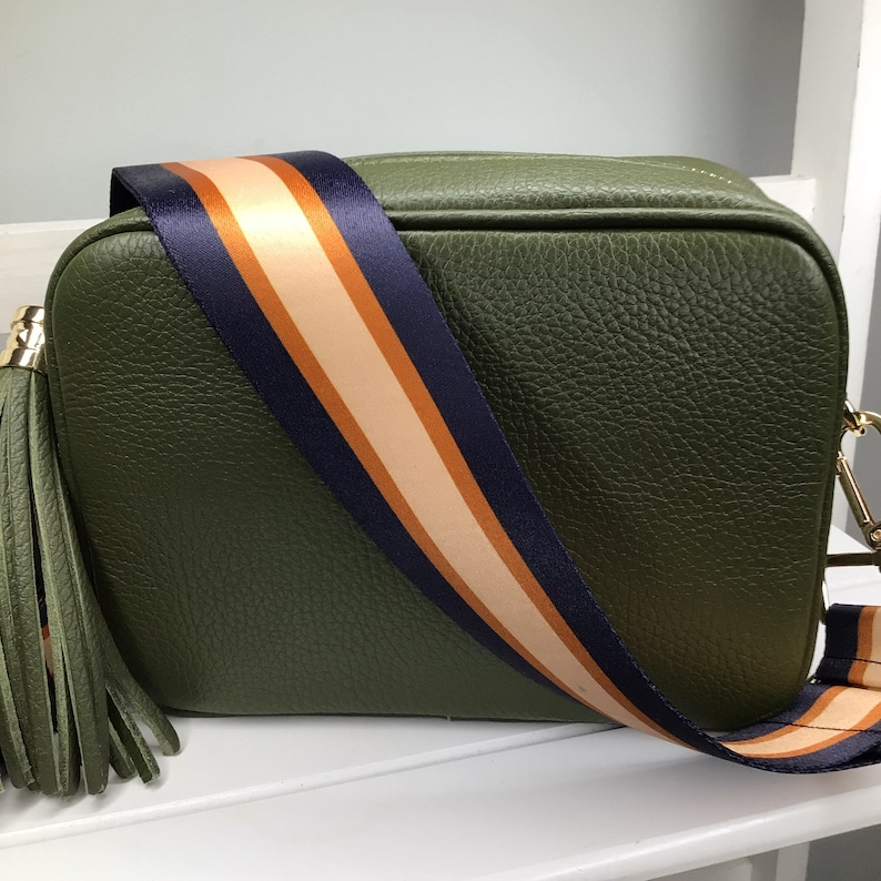 May include: Olive green crossbody bag with a textured finish. The bag features a striped strap in navy, tan, and orange. A matching olive green tassel hangs from the side, and the bag has gold-tone hardware.
