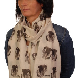 May include: A cream-colored scarf with a black and white border collie dog print. The scarf has a fringed edge and is worn by a person with dark hair.