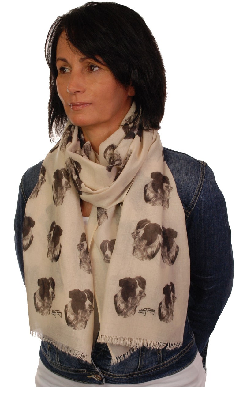 May include: A cream-colored scarf with a black and white border collie dog print. The scarf has a fringed edge and is worn by a person with dark hair.
