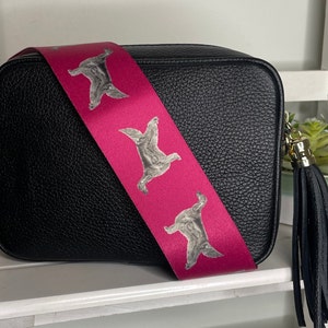 K&ouml;nnte beinhalten: Schwarze Leder-Umh&auml;ngetasche mit einem pinkfarbenen Riemen, der ein sich wiederholendes Muster eines grauen Hundes zeigt. Die Tasche hat einen schwarzen Quasten.