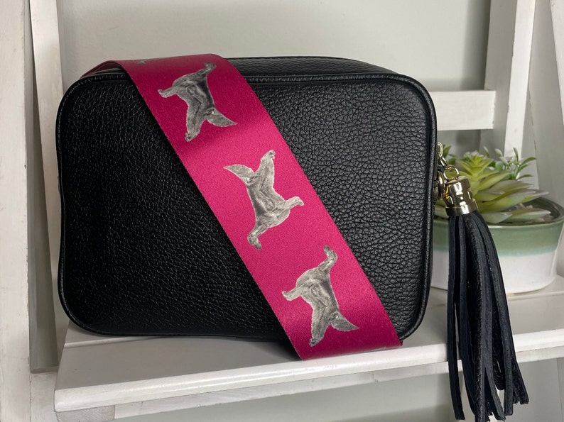 K&ouml;nnte beinhalten: Schwarze Leder-Umh&auml;ngetasche mit einem pinkfarbenen Riemen, der ein sich wiederholendes Muster eines grauen Hundes zeigt. Die Tasche hat einen schwarzen Quasten.