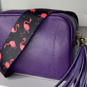 K&ouml;nnte beinhalten: Eine lilafarbene Leder-Umh&auml;ngetasche mit einem schwarzen und pinkfarbenen Flamingo-Musterriemen und einem lilafarbenen Quastenanh&auml;nger.