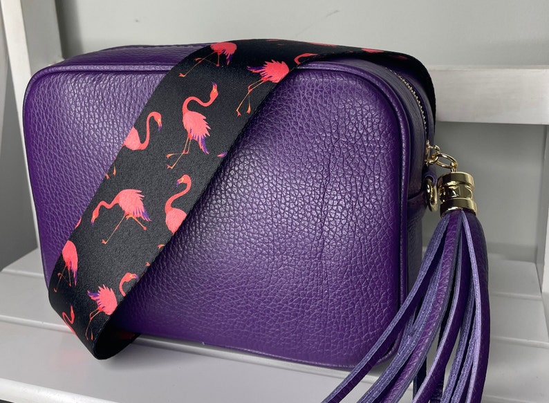 K&ouml;nnte beinhalten: Eine lilafarbene Leder-Umh&auml;ngetasche mit einem schwarzen und pinkfarbenen Flamingo-Musterriemen und einem lilafarbenen Quastenanh&auml;nger.