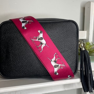 May include: Black leather crossbody bag with a pink strap featuring a repeating pattern of white and brown dogs. The bag has a black tassel.
