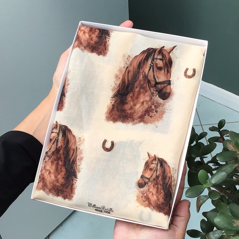 Puede incluir: Una bufanda de seda blanca con un estampado de caballo marr&oacute;n y detalles de herradura dorada. La bufanda est&aacute; en una caja blanca con el texto "William Deville since 1988".